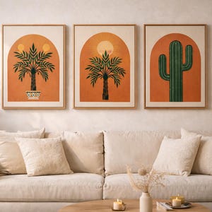 May include: Three framed art prints featuring desert-themed illustrations. The prints depict palm trees and a cactus against an orange background with a sun design. The artwork is displayed above a beige sofa with matching pillows.