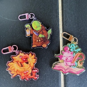 May include: Three acrylic keychains featuring fantasy characters. One keychain depicts a green creature in a brown robe holding a lantern and knife. Another shows an orange bird. The third keychain shows a pink cat-like creature with a quill and open book.