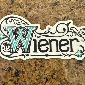 Könnte beinhalten: Ein dekorativer Aufkleber mit dem Wort "Wiener" in einer stilisierten Schriftart. Das "W" ist türkis mit einem schwarzen Filigran-Design. Der Aufkleber hat einen weißen Hintergrund mit schwarzen und türkisfarbenen floralen Akzenten.
