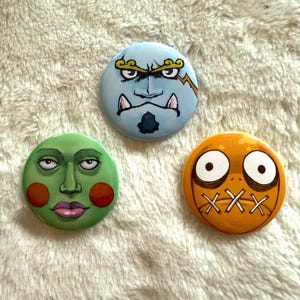 May include: Three round, hand-painted buttons with cartoon faces. One is light blue with an angry face, another is green with a calm face, and the third is orange with a stitched-mouth expression. The buttons are on a white, textured surface.