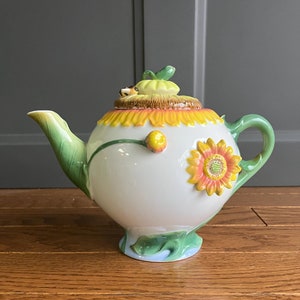 May include: A white ceramic teapot with a sunflower design. The teapot has a green handle and spout, and the lid is shaped like a sunflower.
