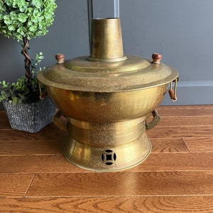 May include: A vintage brass hot pot with wooden handles. The pot has a round base and a tall, cylindrical chimney. The pot is sitting on a wooden floor.