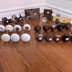 May include: A collection of 14 antique door knobs, including 8 white porcelain knobs and 6 brass knobs. The knobs are arranged in two rows on a wooden floor.