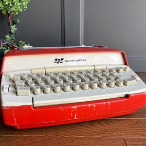 May include: A vintage red and white Smith-Corona typewriter with a white keyboard and black keys. The typewriter has a red plastic case and a white plastic top.