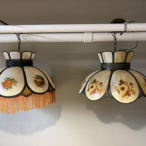 May include: Two hanging pendant lamps with cream-coloured glass panels and dark metal frames. The left lamp features painted fruit motifs and a long orange fringe. The right lamp displays painted yellow floral patterns with a scalloped glass edge.