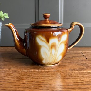 May include: A brown ceramic teapot with a lid, spout, and handle. The teapot has a white floral design on the front. The teapot is on a wooden surface.