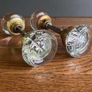 May include: Two antique glass door knobs with brass accents. The knobs have a faceted design and are attached to metal rods.