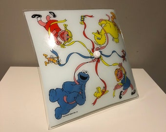 May include: A white ceiling light fixture with a square glass shade featuring Sesame Street characters including Cookie Monster, Big Bird, and a girl with a red dress. The characters are holding colorful ribbons.
