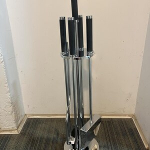 May include: A chrome fireplace tool set with black handles. The set includes a poker, shovel, brush, and tongs, held in a circular stand. The tools have a sleek, modern design, suitable for any hearth. The stand is approximately 20 cm in diameter.