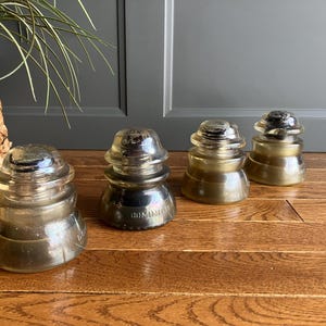 May include: Four vintage glass insulators in various shades of brown and clear. The insulators are stacked on top of each other and have a shiny, glazed finish.