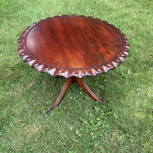 May include: A round, dark brown wooden table with a smooth, polished surface and a decorative, scalloped edge. The table is supported by a central pedestal base that branches out into three curved legs, providing a stable foundation.