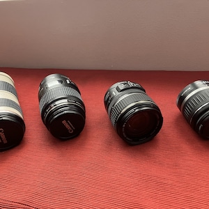 May include: Four Canon camera lenses of different sizes and designs are arranged on a red surface. The lenses are black with silver and white accents. The Canon logo is visible on the lens caps. The lenses are for photography.