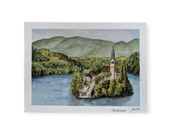 May include: Watercolor painting of a church on an island in a lake, surrounded by trees. The painting features a tall steeple, red-roofed buildings, and green hills in the background. The painting is signed "Slovenia Prita".