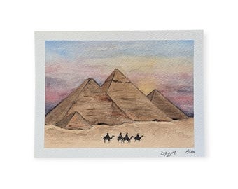 May include: Watercolor painting of the Giza pyramids at sunset. The artwork depicts the pyramids in shades of brown and tan, with a colorful sky in the background. Silhouetted figures on camels are in the foreground. The text "Egypt Bita" is in the lower right corner.