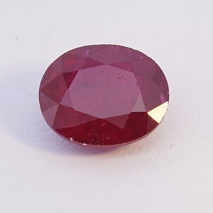 May include: A deep red oval-shaped ruby gemstone with a faceted cut.