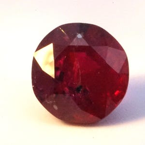 May include: A deep red gemstone with a faceted cut. The gemstone is round and has a high polish.