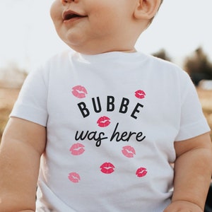 May include: A white baby t-shirt with the text "BUBBE was here" in black lettering and pink lips printed around the text.