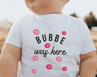 May include: A white baby t-shirt with the text "BUBBE was here" in black lettering and pink lips printed around the text.