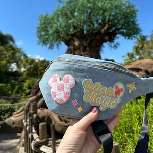 Peut inclure: Une banane en jean bleu clair avec un patch tête de Mickey Mouse rose et blanc, un patch "Believe in Magic" en paillettes dorées et un patch tête de Mickey Mouse avec un ballon rouge.