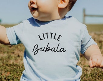 May include: A baby wearing a light blue t-shirt with the text "LITTLE Bubala" in black. The baby is sitting on a patch of green grass.