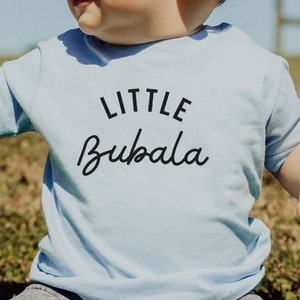 May include: A light blue baby t-shirt with the text "LITTLE BUBALA" printed in black.
