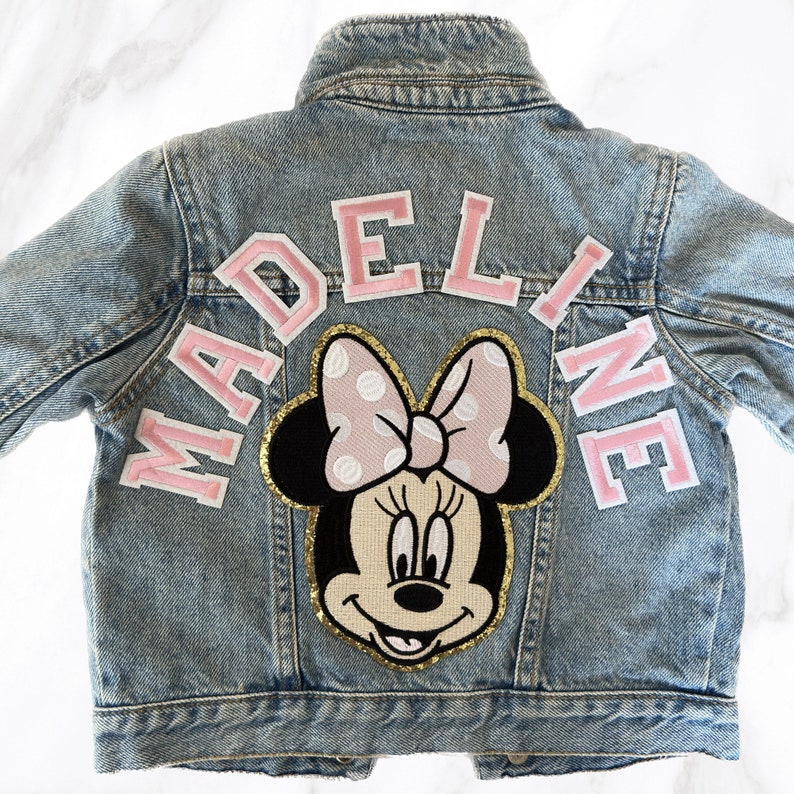 Op de afbeelding: Een lichtblauw denim jack met een roze en witte Minnie Mouse-patch en de naam "MADELINE" in roze letters.