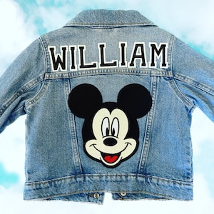 Pode incluir: Uma jaqueta de ganga azul com um remendo do Mickey Mouse e o nome "WILLIAM" em letras pretas nas costas.