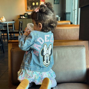 Op de afbeelding: Een jong meisje draagt een blauwe denimjas met een roze en witte "MADEL" patch en een roze en witte Minnie Mouse patch. Ze zit op een bruine leren bank.