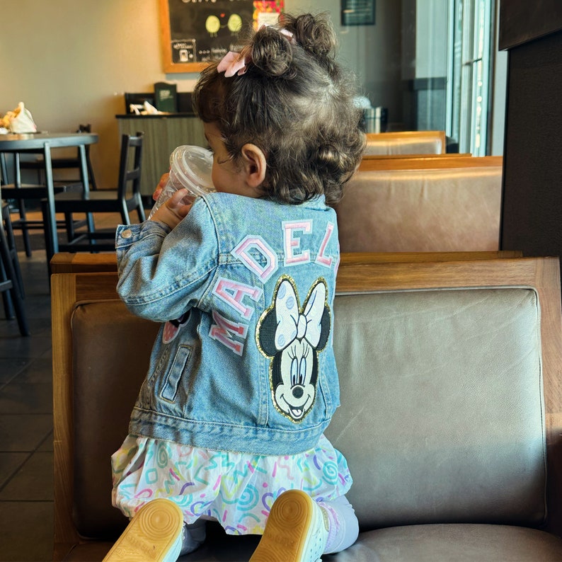 Op de afbeelding: Een jong meisje draagt een blauwe denimjas met een roze en witte "MADEL" patch en een roze en witte Minnie Mouse patch. Ze zit op een bruine leren bank.