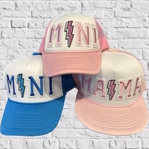 May include: Three trucker hats with white mesh panels and pink, blue, and white brims. The hats have white felt patches with the words "MINI", "MINI", and "MAMA" in pink and blue letters with a pink lightning bolt graphic.