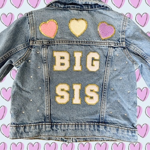 May include: A light blue denim jacket with the words "BIG SIS" in white and gold letters on the back. There are three felt hearts in pink, white, and purple on the top of the jacket.
