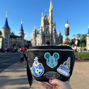 May include: Black fanny pack with a variety of colorful embroidered patches, including Mickey Mouse, Olaf from Frozen, and Cinderella's castle. The patch with Mickey Mouse is green, the Olaf patch is blue, and the Cinderella's castle patch is white with gold accents. The fanny pack also has a red and white patch that says "Come to the Dark Side".