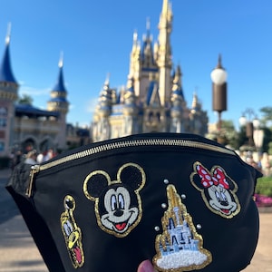 May include: Black fanny pack with gold zipper and embroidered patches of Mickey Mouse, Minnie Mouse, Pluto, and a castle. The background features a castle and blue sky.