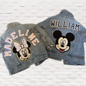 Op de afbeelding: Twee spijkerjacks met gepersonaliseerde patches. Een jack heeft een roze "MADEINE" patch en een Minnie Mouse patch. De andere jack heeft een zwarte "WILLIAM" patch en een Mickey Mouse patch.