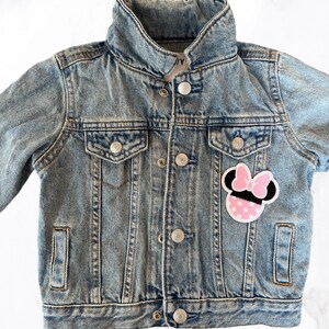 Op de afbeelding: Een lichtblauw denim jack met een roze en zwarte Minnie Mouse-patch op de voorkant.