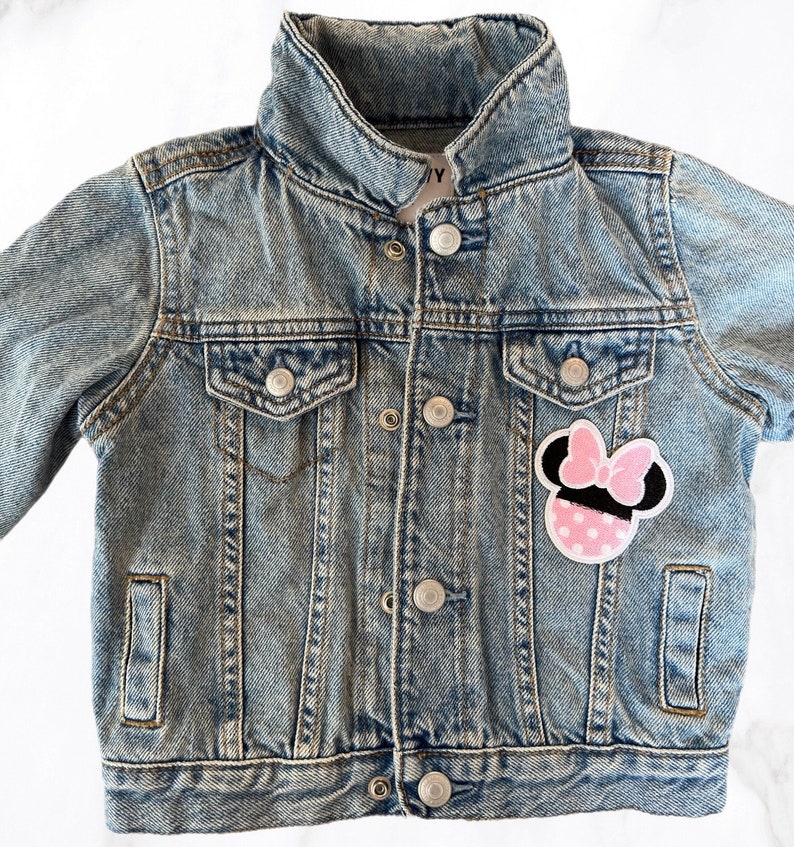 Op de afbeelding: Een lichtblauw denim jack met een roze en zwarte Minnie Mouse-patch op de voorkant.