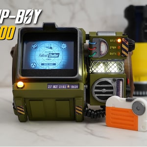 Op de afbeelding: Een gedetailleerde Pip-Boy 2000 replica uit de Fallout-serie. Het olijfgroene apparaat heeft een scherm met "Fallout Shelter" en diverse knoppen en lampjes. Een oranje en wit apparaat is aan de zijkant bevestigd.