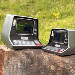 Può includere: Due terminali per computer in stile retrò con custodie grigie e nere, tastiere e piccoli schermi. Gli schermi mostrano testo verde. I terminali sono su una roccia con uno sfondo di erba verde. Il testo "RadCat" è visibile sulla tastiera.