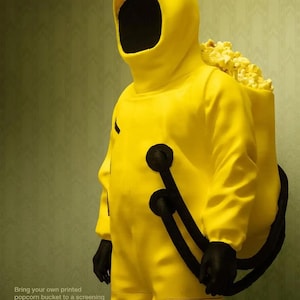 Peut inclure: Image promotionnelle pour le film "Backrooms" montrant une personne dans une combinaison jaune vif avec un sac à dos rempli de pop-corn. La combinaison a des gants noirs et une capuche avec un vide sombre à la place du visage. Le texte "Backrooms 05.29.26" est en haut.