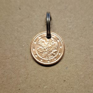 Könnte beinhalten: Eine kupferfarbene Münze mit einem silbernen Ring, der an einem silbernen Schlüsselring befestigt ist. Die Münze zeigt ein Eichenblatt-Design und das Jahr 2004.