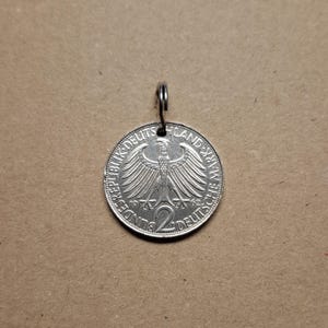 Könnte beinhalten: Eine silberne deutsche 2-Deutsche-Mark-Münze mit einem Loch, das oben zum Aufhängen gebohrt wurde. Die Münze zeigt den deutschen Bundesadler und den Text "Bundesrepublik Deutschland" und "Land Berlin".