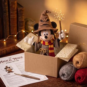 May include: A crocheted Mickey Mouse figure wearing a brown wizard hat and a red and yellow striped scarf, holding a glowing wand. The figure is inside a cardboard box with yarn balls, a crochet hook, and a printed pattern titled The Magical Mouse.