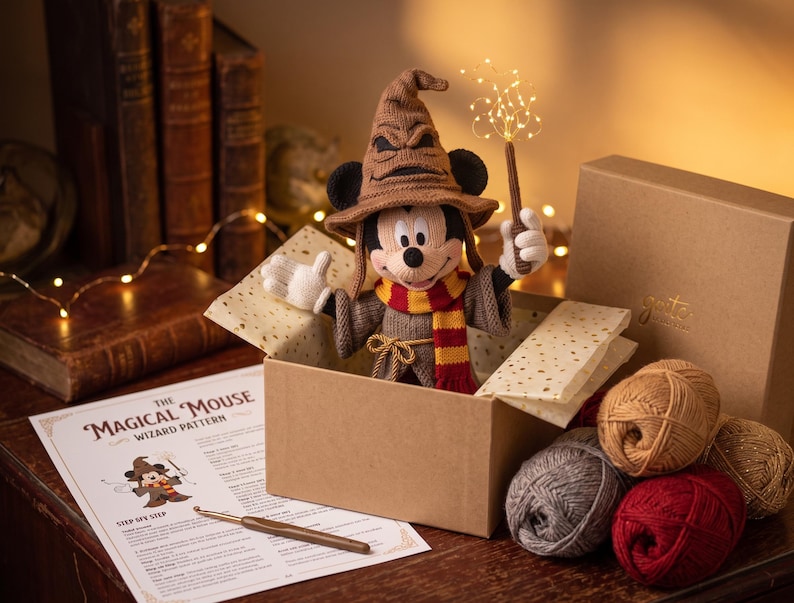 May include: A crocheted Mickey Mouse figure wearing a brown wizard hat and a red and yellow striped scarf, holding a glowing wand. The figure is inside a cardboard box with yarn balls, a crochet hook, and a printed pattern titled The Magical Mouse.