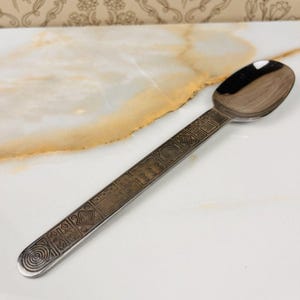 May include: A silver spoon with an engraved handle featuring geometric patterns. The spoon's bowl is polished and reflective. The handle's design includes squares, circles, and diamond shapes. The spoon is set against a white and gold marble-like surface.