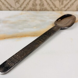 May include: A silver spoon with an oval bowl and a long handle. The handle is decorated with geometric patterns, including circles, squares, and lines. The spoon is made of metal and has a shiny finish.