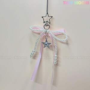 May include: A pink ribbon with a pearl and star charm. The charm is a silver star with a clear rhinestone center. The ribbon is tied in a bow and hangs from a silver star-shaped hook.