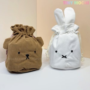 May include: Two drawstring bags, one brown bear-shaped and the other white bunny-shaped. Both are made of corduroy fabric. The bear bag has embroidered eyes and a nose, while the bunny bag has embroidered eyes and a mouth.