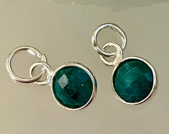 Pode incluir: Dois pingentes prateados com pedras preciosas redondas, facetadas, verde-azulado. Cada pingente tem um pequeno laço na parte superior para prender a uma corrente ou pulseira. As pedras são fixadas em uma moldura prateada.