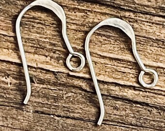 May include: Two silver-colored earring hooks are displayed against a textured, brown wooden surface. The hooks have a simple, curved design with a small loop at the bottom for attaching earrings. The hooks are made of a shiny metal.