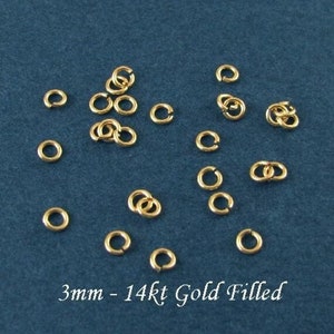 May include: A close-up of 17 gold-filled jump rings, each measuring 3mm in diameter. The rings are arranged in a scattered pattern on a blue surface. 3mm - 14kt Gold Filled